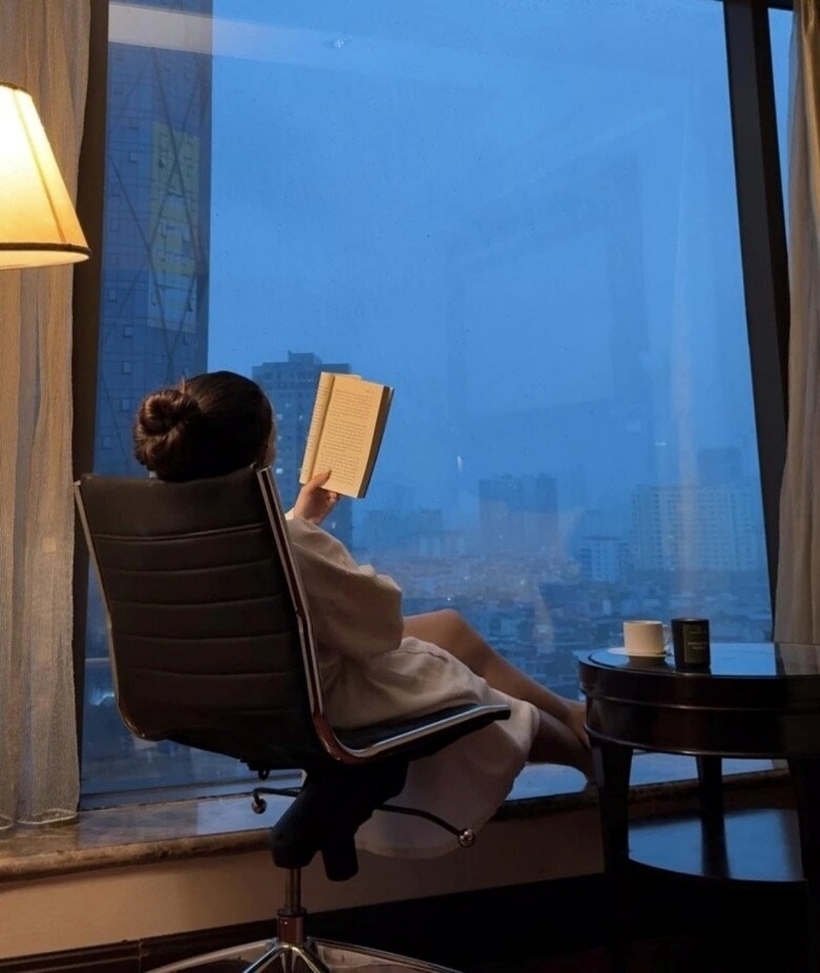 Yến Xuân đọc sách trong phòng ngủ có cửa kính rộng, view thành phố.